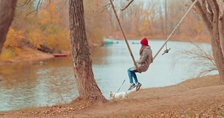 Small dog is standing by the river, and the mistress is swinging on a swing. Girl holds a puppy on a leash, walk.