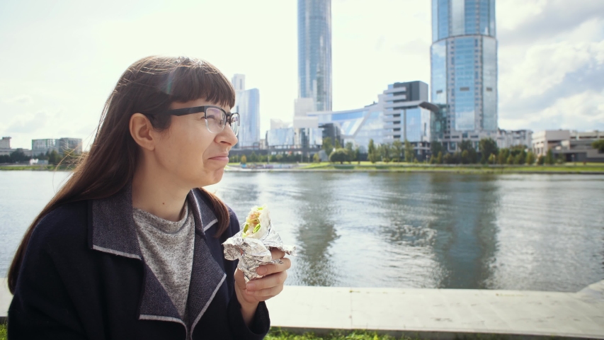 Happy businesswoman in glasses eats doner kebab lunch. Girl sitting on concrete fence near riverside in downtown.