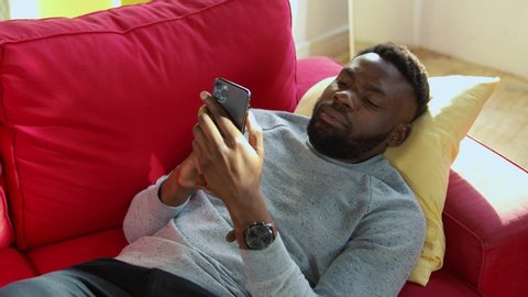 Young attentive african man getting up on couch busy reading content on smartphone checking up news staying at home.