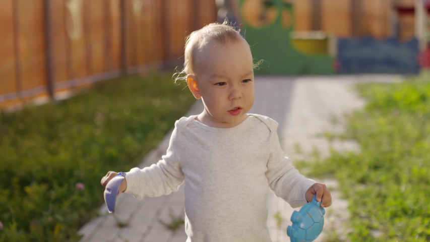 Tilting shot of Caucasian toddler in bodysuit and sandals walking on footpath in garden, carrying toy sand shovel and plastic mold and putting it in his mouth