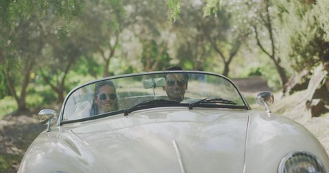 Happy smiling diverse newly wed couple driving on a sunny country road, bride and groom driving together in a white vintage convertible, bride holding flower bouquet