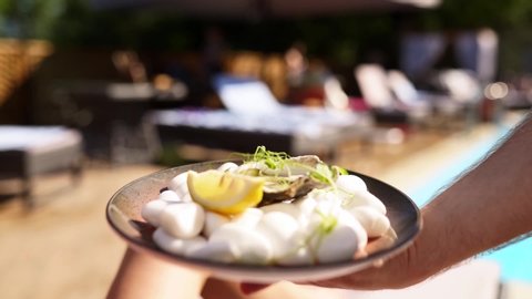 Waiter carries oysters and scallops on tray, with slices of lemon with a swimming pool on background at luxury hotel or beach club. Man serving food in slow motion at resort. Seafood on a plate.