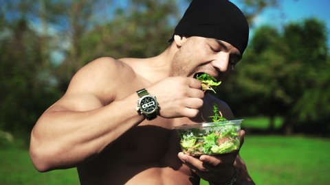 Young muscular sportsman eating healthy food outdoors and looking on camera. Fresh salad and carrot are rich in vitamins needed for developing strong muscular tissue. Naked sexy athlete preparing for