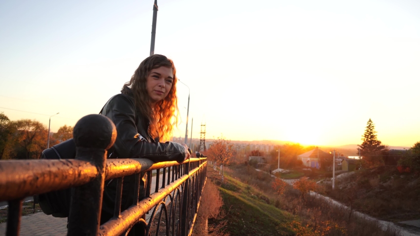 A young beautiful woman in a leather jacket with a black backpack stands thoughtfully on the bridge against the background of the urban district.