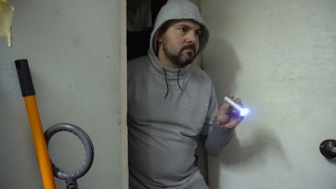 Bearded man in a hood with a flashlight in his hand. Dressed in a gray sweatshirt. Enters the door. Robbery. Professional thief. Maniac in the house. Angry facial expression.