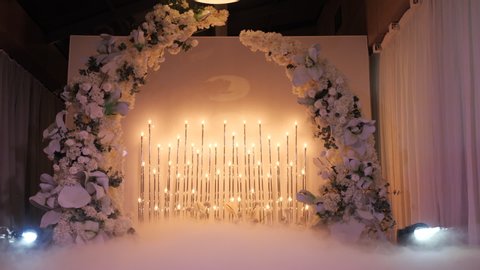 Illuminated wedding flower arch before the wedding night ceremony. Floral decor in the restaurant hall. Atmosphere, flashlights and fog from a smoke machine, light spotlights.