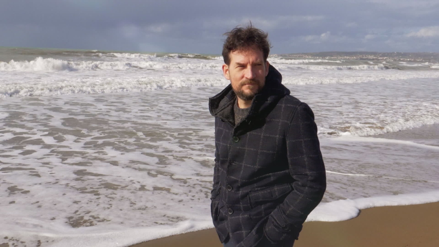 Happy young man with beard walking in front of the sea with wild waves in autumn slow motion