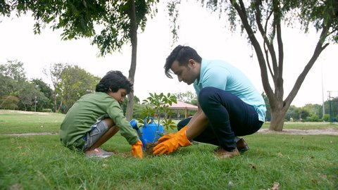 A young attractive man gardening a little sapling with his son in a park - ecology concept. Cute little boy helping his father in planting the tree while working together in a garden - Go green con...