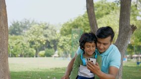 Young father and his son waving a 'Hi' while having a conversation on a video call - technology concept. Cute Indian child sitting with his dad in a park and happily talking to his mother over a vi...