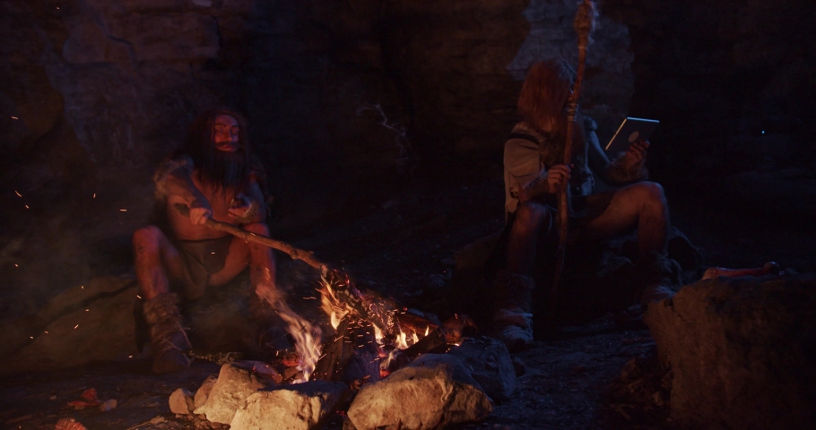 Slow motion tribe cavemen neanderthals wearing animal skin use tablet computer sitting by the fire in a cave 