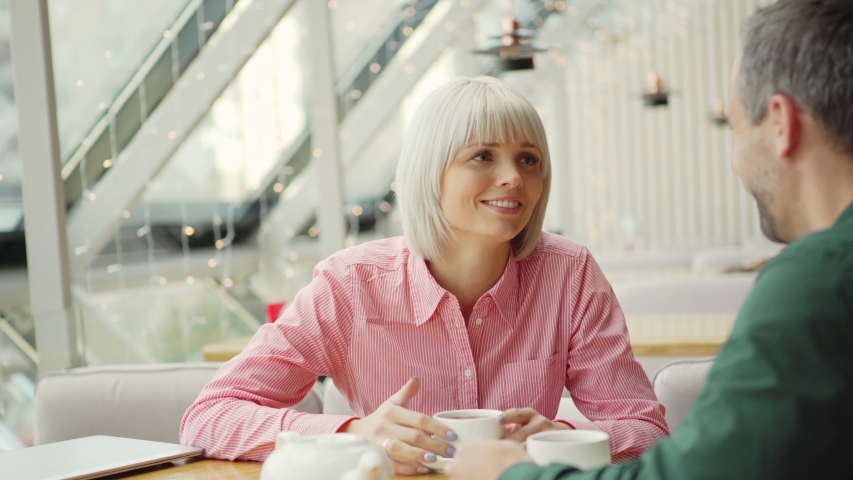 Happy female with cup of hot drink smiling and telling story to adult male while sitting at cafe tale during romantic date
