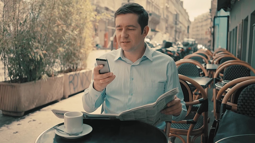 Businessman drinks coffee at lunch, reads a newspaper and speaks on the phone in Paris cafe.