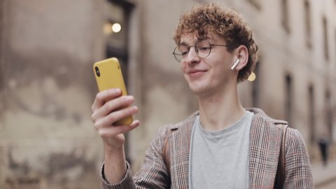 Close Up of Millennial in Earphones and Glasses Video Chatting with Friends, Talking, Laughing via Modern Mobile Phone, Walking at Old City Street Background. Communication Concept.