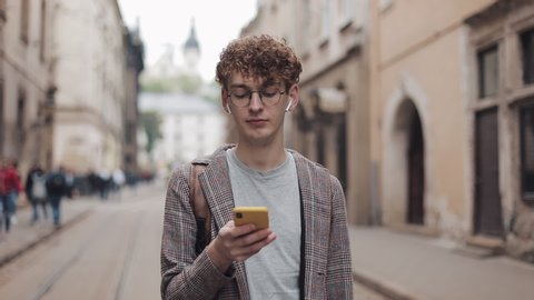 Portrait of Young Guy in Glasses and Earphones Wearing Jacket, Holding Smartphone and Looking to Screen than to Camera at Old City Background.