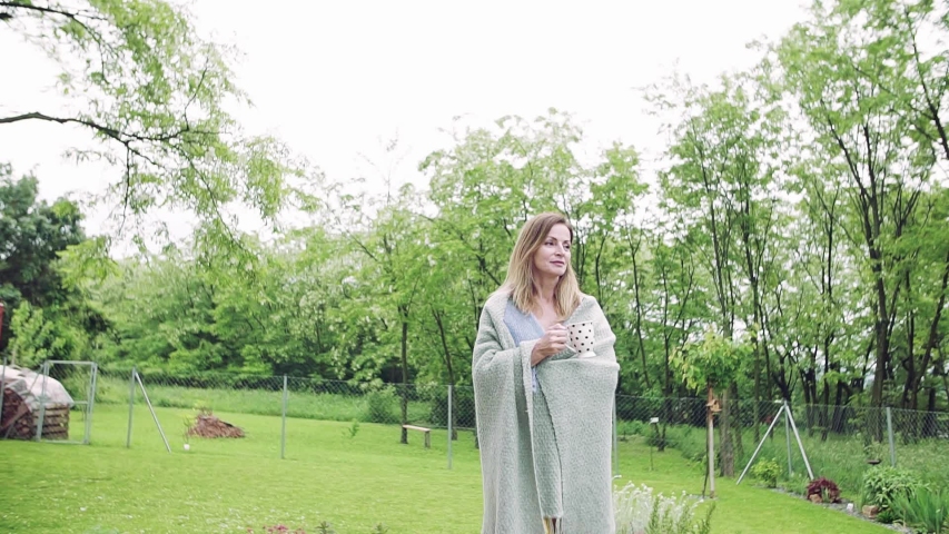 A mature woman with tea and blanket walking outdoors in the garden.
