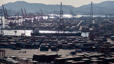 Timelapse Busan harbour cargo port with containers ready for shipment and operating cranes with water reflecting sunlight against hill silhouettes in early morning zoom in