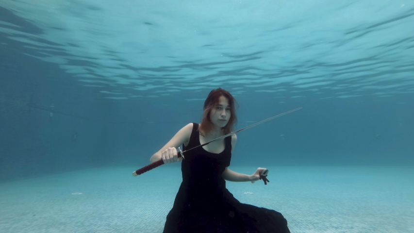 Unusual girl sitting underwater at the bottom of the pool in a black dress with a red samurai sword in his hand. She looks at him and plays with him. Fashion portrait. Concept. Slow motion. 4K.