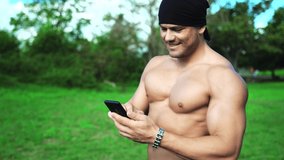Muscular build fitness trainer walking in the park and chatting online with clients using mobile phone. Sportsman with perfect torso using modern sport mobile apps and sending workout instructions to