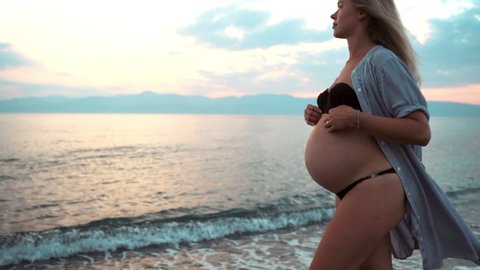 Attractive blonde woman in swimsuit walking in slow motion along the sea beach in amazing pink sunset. Woman's hands embracing her belly, love and tenderness of pregnant woman