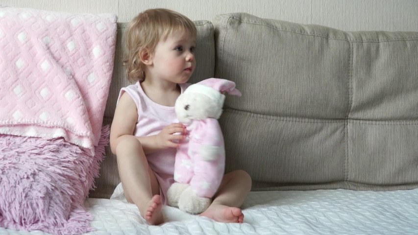 Portrait of little smiling cute 1 year old baby girl plays teddy bear pink toy, hugs, kisses and laughs. Funny child after sleep rejoices his toy
