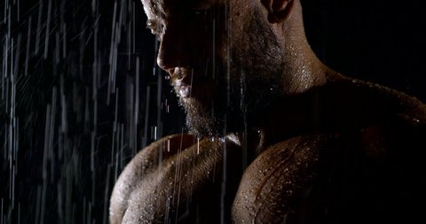 The face of a brutal bearded muscular male bodybuilder close-up on a black background, he is in the rain, water flows down him.
