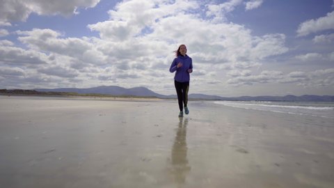Low dolly shot of a girl running, jogging on the shore of a sandy beach with Atlantic ocean waves on a wonderful sunny day in Ireland in 4K