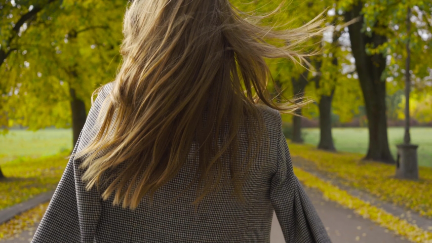 Back view of a smiling Caucasian woman in checkered jacket walking along the road. Young brunette girl strolling in the autumn park.