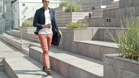 Handheld Wide Shot Of Handsome Guy With Coffee Cup And Bag Walking To Sit Down On Steps