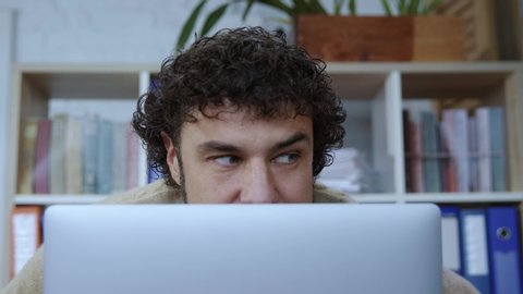 Funny caucasian man with curly hair hiding head behind monitor spying his colleagues working by computer in corporate office. Close-up.
