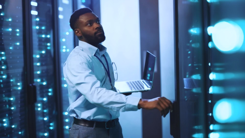 Afro-american IT administrator opening racks of server computers running diagnostics on laptop technology monitoring system of data center. Slow motion. | Shutterstock HD Video #1039586453
