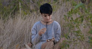 Attractive short-haired Caucasian hipster girl sitting in the dry grass and putting on the earphones, searching and listening her favorite music songs in her smartphone slow motion 4k 50 fps