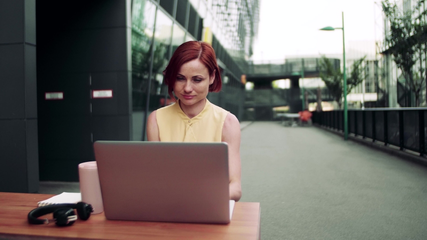 Young businesswoman with laptop sitting in cafe outdoors in city, working.