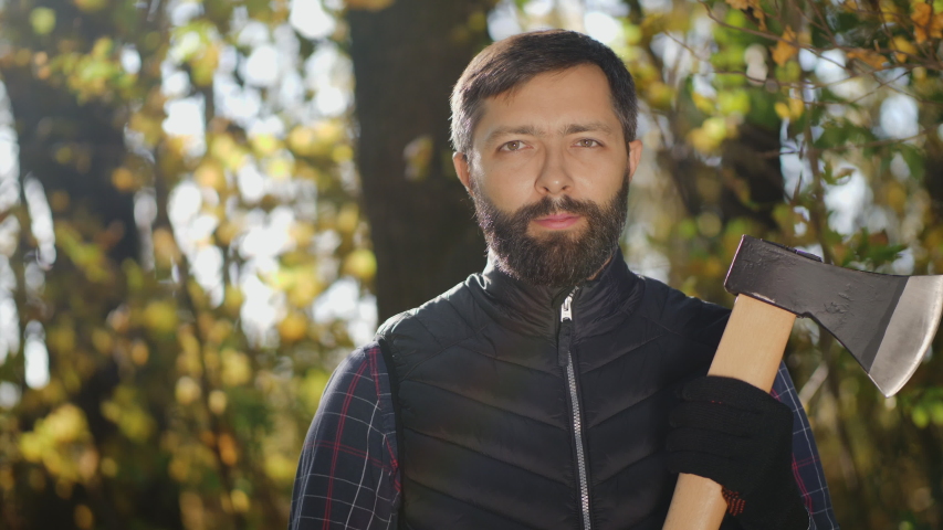 Lumberjack standing with his ax in the woods, looking at camera and smile. Portrait of a handsome bearded man with an ax