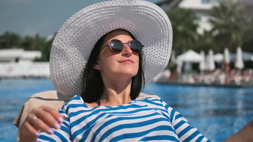 Relaxed smiling woman in sunglasses and white hat sunbathing near swimming pool medium close-up