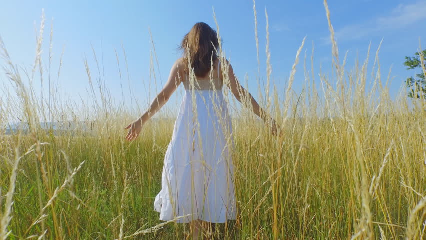 Beautiful young woman with a long white dress walking through long grass and gently touching the stems.