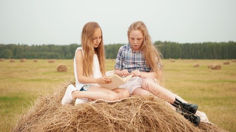 Teenager girls reading book on haystack at countryside field at vacation. Surprised girls looking book on hay stack at harvesting field in village
