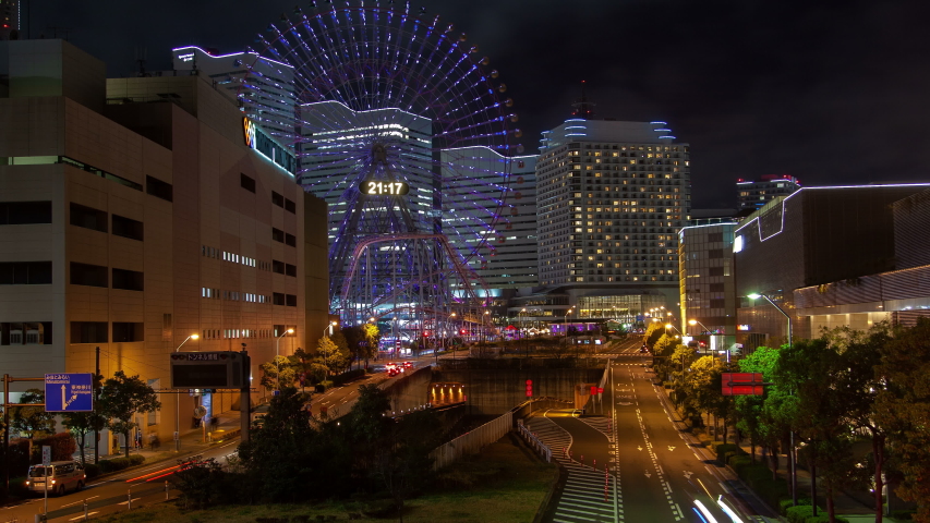 Timelapse wide Tokyo road with traffic past green trees by modern buildings and large illuminated Ferris wheel under cloudy sky at night zoom out