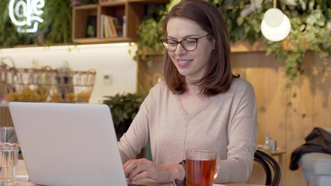 Enthusiastic smart woman in glasses typing on laptop and drinking tea at table in cute cafe