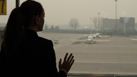 Thoughtful woman stay at window, with arm on glass. Passenger looking out to small bizjet plane parked at apron of large international airport. Silhouetted portrait shot from back