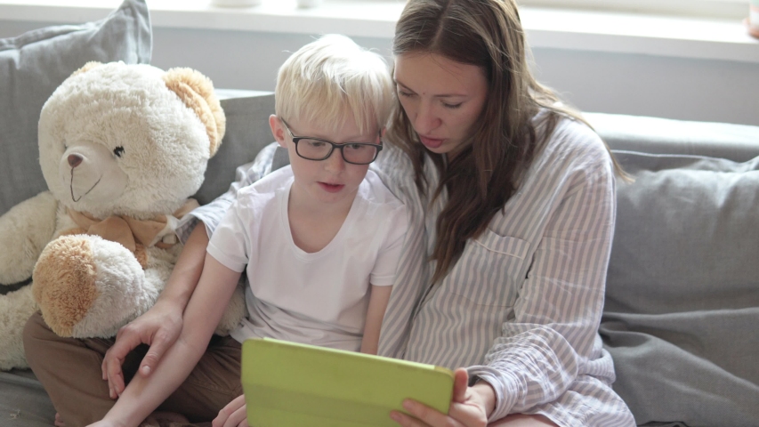 Mom and son are sitting on the couch and calling dad on the tablet. Woman and child skype relatives