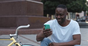 Excited African American Guy in Headphones Using his Smartphone Listening to Music Choosing Song Smiling while Sitting on Steps near Bike at the City Background. Close up