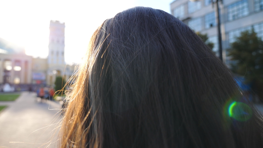 Unrecognizable woman going to work and straightening hair at early morning street. Pretty girl having stroll through city street on way to job. Sunlight illuminating hair of charming brunette. Slow mo