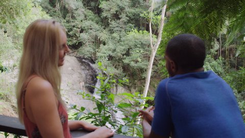 Interracial friends talking in front of small waterfall in an Australian rainforest by safety railing during daytime. Medium to closeup shot with camera zoom out on 4k RED camera.