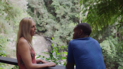 Interracial friends talking in front of small waterfall in an Australian rainforest by safety railing during daytime. Medium to closeup shot with camera zoom out on 4k RED camera.