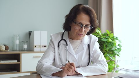 60s American female doctor talking and writing, working at table in clinic office interior. 4k Portrait of serious mature expert speaking and doing paperwork sitting in interior. Concept: healthcare
