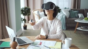 American old businesswoman using vr headset working at desk with laptop in office room. Elderly female testing virtual game and digital innovation with mask on face. Elegant 60s lady sitting at modern