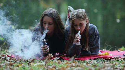 Vape lgbt teenagers. Bisexual lesbian young caucasian teenage girls in casual clothes vape electronic cigarette near the river on the street in the park in the autumn evening. Bad habit.