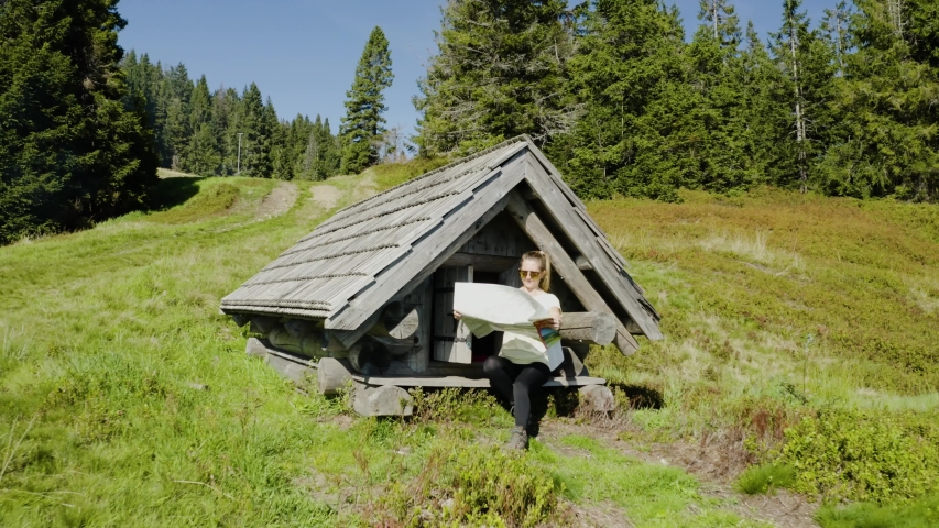 Young woman reads unfolded map, sitting in front of small old wooden mountain hut, placed in the middle of mountain clearing. Drone flight back