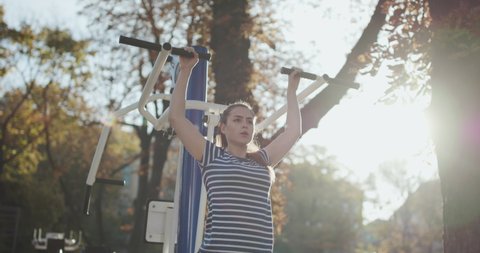 Strong fit Caucasian woman doing pull ups in morning workout in park. Strong muscular woman does chin ups on metal bar. Young brunette doing chin-ups on the gym bar outdoorsの動画素材