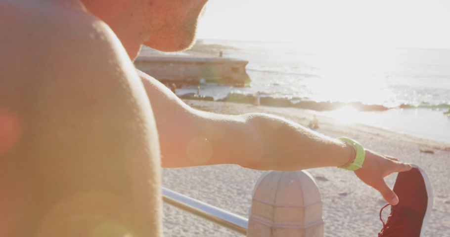 Front view close up of a young Caucasian man in sportswear stretching his legs on a fence before a run on a sunny day by the sea